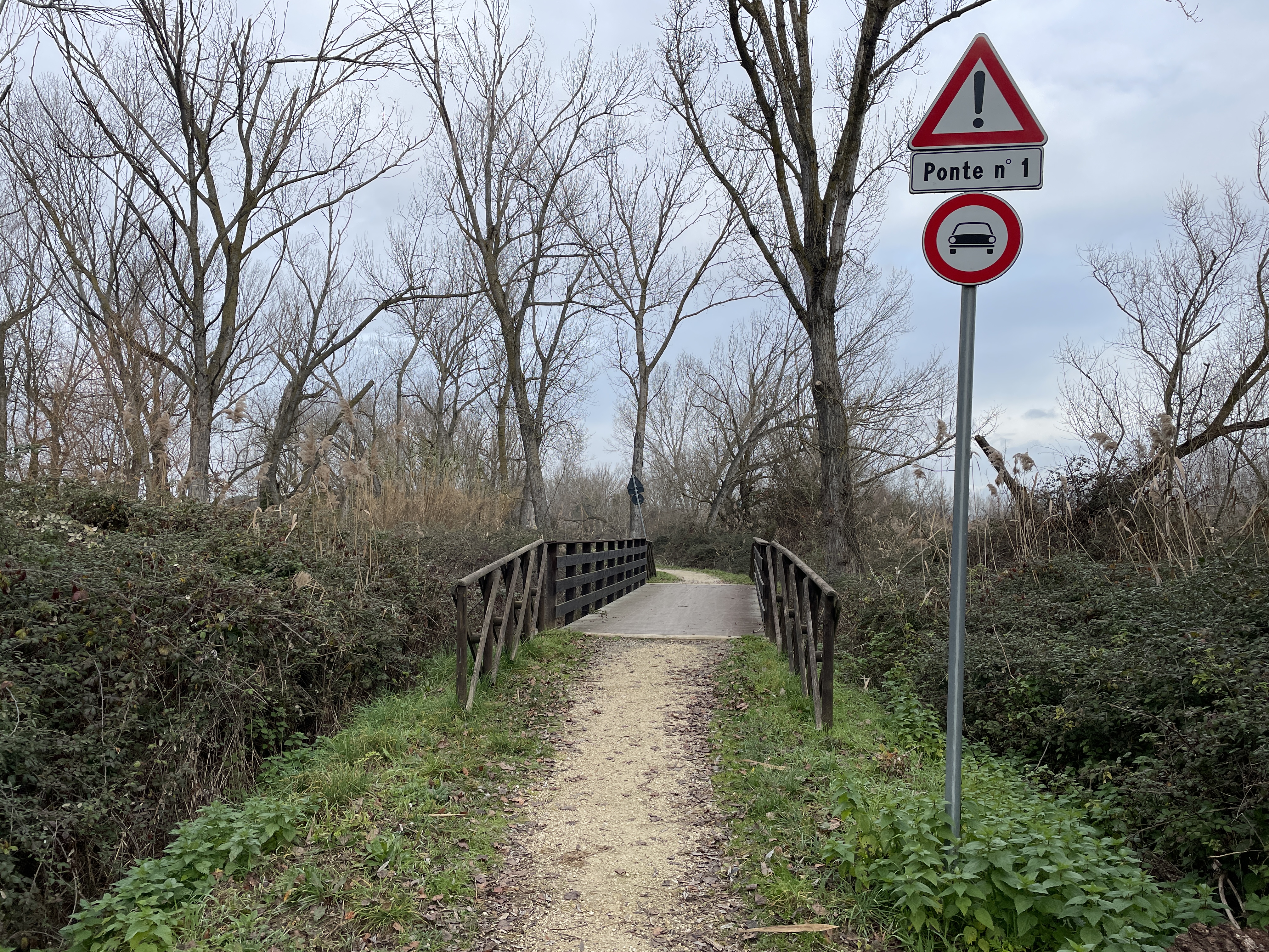 Schotterweg zur Holzbrücke mit Holzgeländer. Zuerst, auf der rechten Seite, Straßenschilder ohne Zugang für Autos